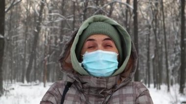 Koruyucu tıbbi maske takan mutlu kadın, kışın bir şehir parkındaki kar yağışı altında ağaçların arkasında seviniyor. Pozitif kadın kışın sevinir ve beyaz kar yağar.