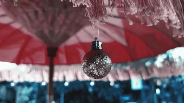 Primer plano de la bola de plata gris colgando de la franja de sombrilla roja al aire libre. 4k. Decoración de Navidad en el resort tropical en diciembre. — Vídeos de Stock
