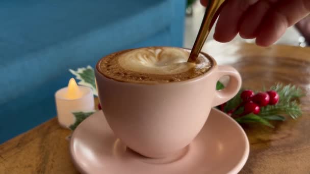 Mão mexendo café com leite latte em uma caneca rosa na mesa decorada com ramo de árvore de natal e vela LED com grão de filme — Vídeo de Stock