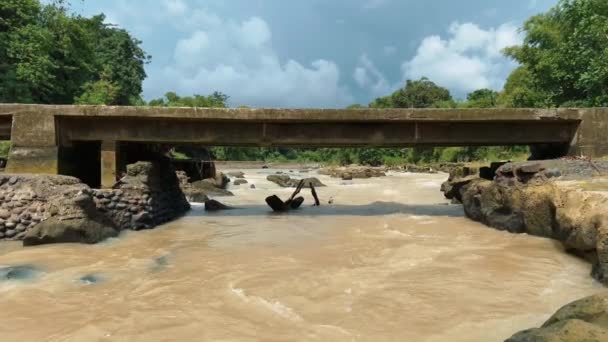 Kahverengi çamurlu bir nehrin üzerinde beton bir köprü. Bali adası manzarası. Volkanik taş kıyısı ile nehir akışı. — Stok video