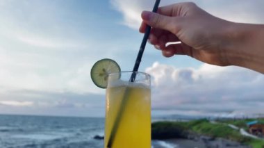 Woman stirring tropical cocktail with lime slice on glass. Seaside bar view. POV enjoy vacations at ocean shore.