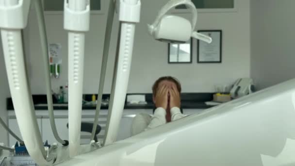 Грустный и испуганный человек, прячущий лицо в руках, сидя в кресле у дантистов. Процедура жестких зубов в белом медицинском кабинете. — стоковое видео