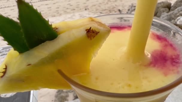 Makro taze mango ve ananas suyunu beyaz kumsalda kızılcık şurubuyla karıştırıyor. Plaj barında bir bardak kokteyle tropik meyve dilimi. — Stok video