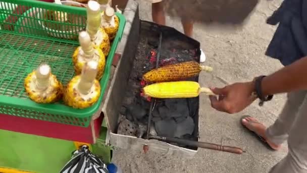 Delicious street food on a beach. Man waving fan and turning around corn on grill. Cafe on wheels at seaside. Trolley with Barbecue corn close-up. — Stock Video