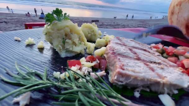 Zbliżenie materiału z grillowanego tuńczyka i puree ziemniaczanego z sałatką na plaży. Ręka z fok jedząc talerz ryb z warzywami i rozmarynem w kawiarni na plaży. — Wideo stockowe