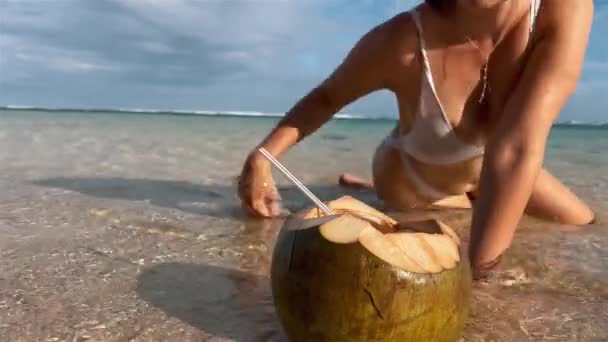 Une jolie fille en maillot de bain blanc arrive à une jeune boisson à la noix de coco pour s'allonger sur du sable blanc dans de l'eau cristalline de l'océan et siroter un cocktail coco profitant d'une plage sauvage dans un paradis tropical. — Video
