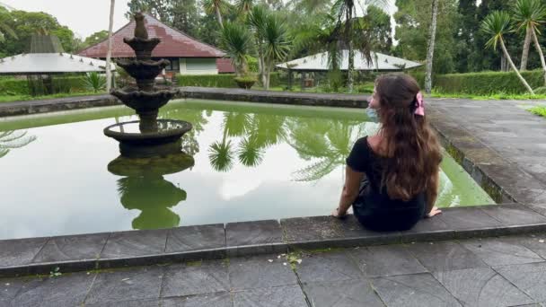 Chica con lazo rosa y máscara médica sentada al lado de la piscina de una fuente rota en el jardín botánico de Bali. Tiempo libre pandémico al aire libre. — Vídeo de stock
