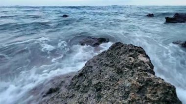 Okyanus suyunun resif kayalarına sıçrayışının yakın çekim görüntüleri. Akşamüstü mavi saatte beyaz köpük plaj taşlarını yıkıyor..