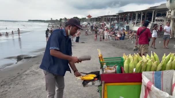 Bali, Indonésie - 28. ledna 2022: Pouliční kuchař mává větrákem nad grilem se žlutými kukuřicemi. Proces tvorby pouliční stravy na pláži s kavárnami a lidmi.