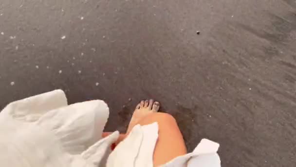 POV syn på en flicka bär vitt linne klänning bär söta skor med rosetter i handen trampar på seacomb med svart sand och vitt havsskum. — Stockvideo