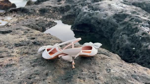 Hermosos zapatos femeninos blancos con tacones altos y lazo en un arrecife del océano negro a la orilla del mar al atardecer. Calzado femenino en una roca en la orilla del océano. — Vídeo de stock