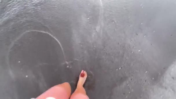 黒と灰色の砂の上を歩く茶色の花とピンクの脚とフリップフロップを身に着けている女性のポルノビュー海の水は男性サーファーを反映. — ストック動画