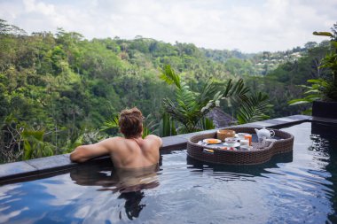 Anonim bir adam havuz kenarında takılıp, ormanın derinliklerine bakarken yüzen bir kahvaltı tepsisi yüzme havuzunun su yüzeyinde yüzüyor..