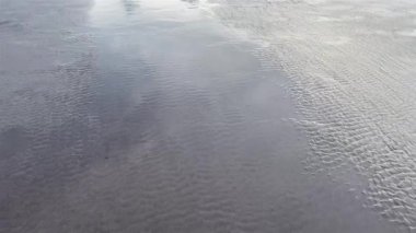 Suyu mavi gökyüzünü ve bulutları yansıtan siyah kumsal. Köpük banyosuyla deniz tarağına basıyorum..