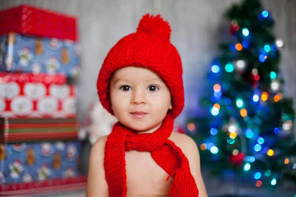 Μικρό αγόρι με κόκκινο καπέλο και κασκόλ, μπροστά από ένα χριστουγεννιάτικο δέντρο — Φωτογραφία Αρχείου