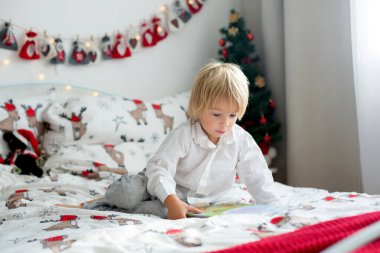 Güzel bir çocuk, Noel 'de yatakta kitap okuyor, etrafında süslemeler yapıyor.