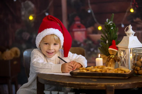 かわいい幼児の子供 クリスマスの男の子 サンタクロースへの手紙を書いて家でクッキーを食べる 居心地の良い木製のコテージ — ストック写真