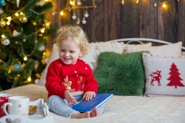 Tatlı bebek, Noel kıyafetli kıvırcık sarışın kız, Noel 'de ahşap bir kulübede oynuyor, etrafında süslemeler yapıyor. Çocuk kitabı okuyor ve çay içiyor.