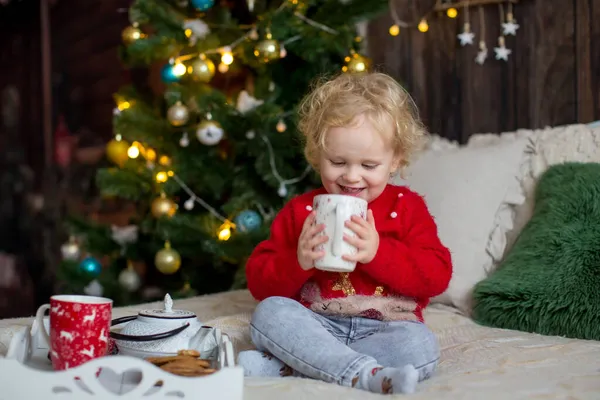 かわいい幼児の子供 クリスマスの服の巻きブロンドの女の子 クリスマスに木製のキャビンで遊んで 彼女の周りの装飾 児童書やお茶を飲む — ストック写真