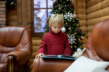 Sarışın çocuk, kitap okuyor ve Noel zamanı, dağlarda rahat bir kulübede hediyeler tutuyor.
