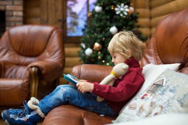 Sarışın çocuk, kitap okuyor ve Noel zamanı, dağlarda rahat bir kulübede hediyeler tutuyor.