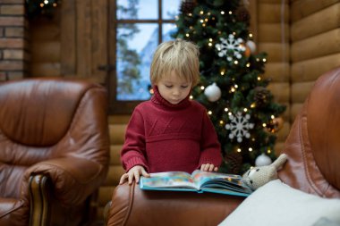 Sarışın çocuk, kitap okuyor ve Noel zamanı, dağlarda rahat bir kulübede hediyeler tutuyor.