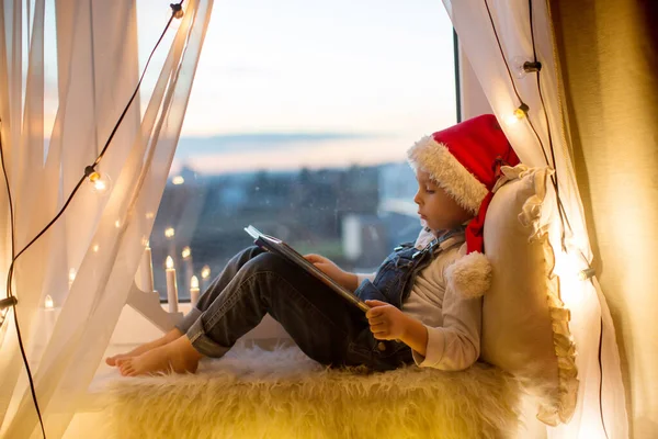 蹒跚学步的孩子 可爱的金发男孩 坐在窗上看书 圣诞节的灯光环绕着他 — 图库照片