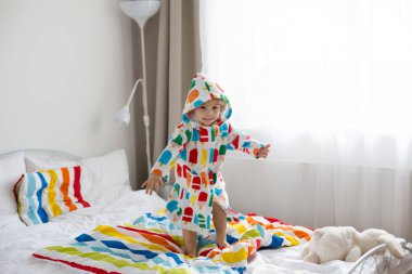 Mutlu çocuk, renkli bornozlu sarışın çocuk, yatağa atlıyor, mutlu bir şekilde gülümsüyor.