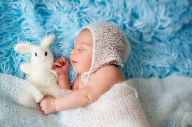 Küçük tatlı yeni doğmuş bebek, beyaz naylona sarılı uyuyor, küçük örgü bir oyuncak tutuyor.
