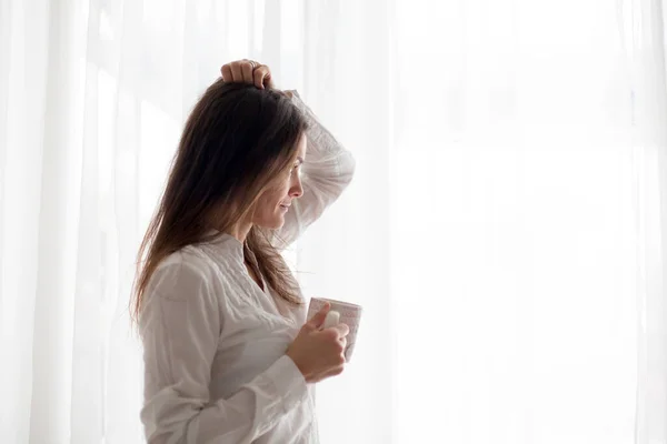 白いドレスを着た中年女性家でコーヒーを飲み窓から外を見る白い背景 — ストック写真