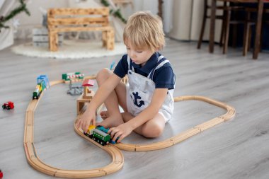 Sevimli sarışın çocuk, evdeki renkli trenlerle ve demiryollarıyla oynuyor.