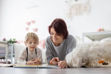 Sevimli sarışın anaokulu çocuğu, evcil köpekli sarışın çocuk, evde annesiyle kitap okuyor.