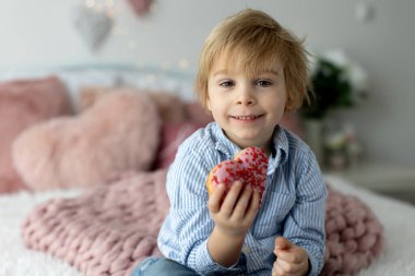 Sevimli küçük sarışın çocuk, anaokulu çocuğu, kalp şeklinde pembe donut yiyor, Sevgililer Günü için yapılmış, Sevgililer Günü geçmişi