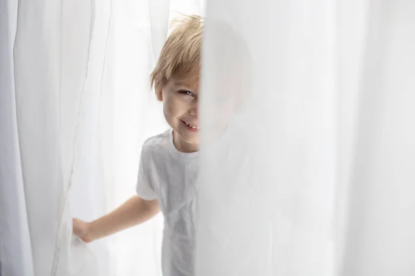 かわいい幸せな幼児の男の子 白い布の子供 カメラで喜んで笑顔 白いカーテンで窓の横に遊んで — ストック写真