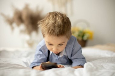 Sarışın anaokulu çocuğu, tatlı çocuk, güneşli bir günde cep telefonuyla yatakta oynuyor.
