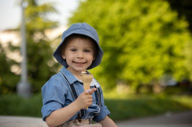 Cute toddler child, eating ice cream outdoors in the park, spring flowers around him, fountain in garden