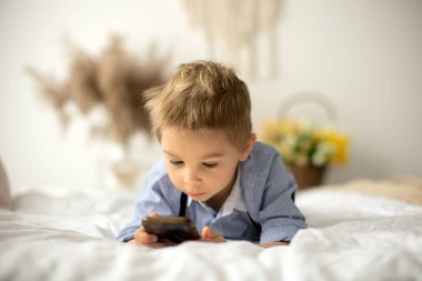 Sarışın anaokulu çocuğu, tatlı çocuk, güneşli bir günde cep telefonuyla yatakta oynuyor.