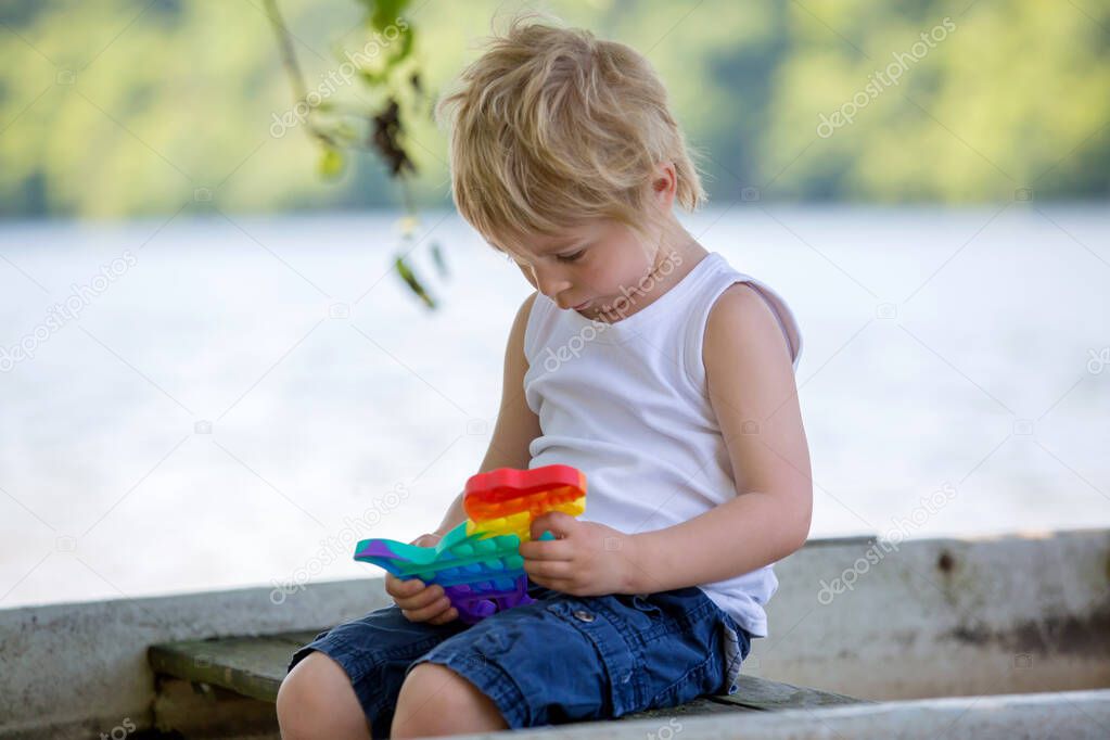 Little toddler child, blond boy, playing with pop it antistress toy in the park in a boat in lake