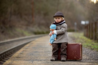 Tren istasyonunda elinde bavul ve oyuncak ayıyla bekleyen sevimli çocuk.