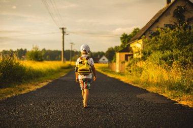 Beautiful child, boy with backpak walking on a little rural path on sunset, after rain, summertime