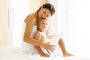 Cute blond toddler child with white tower around belly, sitting in bed with mother after bath, summertime