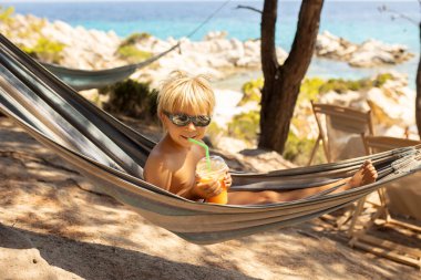 Little boy, drinking coctail, orange juice on the beack in hammock, enjoying summer. Halkidiki, Greece