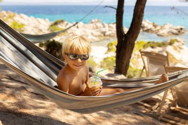 Little boy, drinking coctail, orange juice on the beack in hammock, enjoying summer. Halkidiki, Greece