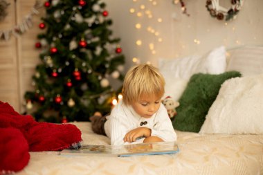 Tatlı sarışın anaokulu çocuğu, kitap okuyor ve Noel 'de rahat evde hediyesini açıyor, ışıklar ve süslemeler.