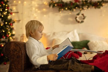 Tatlı sarışın anaokulu çocuğu, kitap okuyor ve Noel 'de rahat evde hediyesini açıyor, ışıklar ve süslemeler.