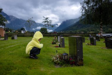 Sad little child, blond boy, standing in the rain on cemetery, sad person, mourning