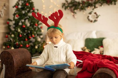 Tatlı sarışın anaokulu çocuğu, kitap okuyor ve Noel 'de rahat evde hediyesini açıyor, ışıklar ve süslemeler.