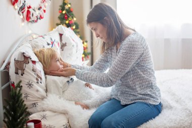 Annesi ateşi kontrol ediyor. Sarışın bebeği, hasta oğlu, Noel 'de yatakta yatıyor, öksürüyor ve burnu akıyor.