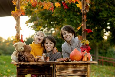 Şirin sarışın çocuk ve kardeş kardeşler, sonbahar ahşabının yanında duruyorlar, süslemeleri, elmaları, yaprakları, kupaları, parkta, sonbahar zamanı
