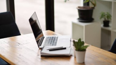 Portatif bilgisayarın yan görüntüsü ahşap masanın üzerinde modern ve rahat ofisin defter ve dekoruyla duruyor. 